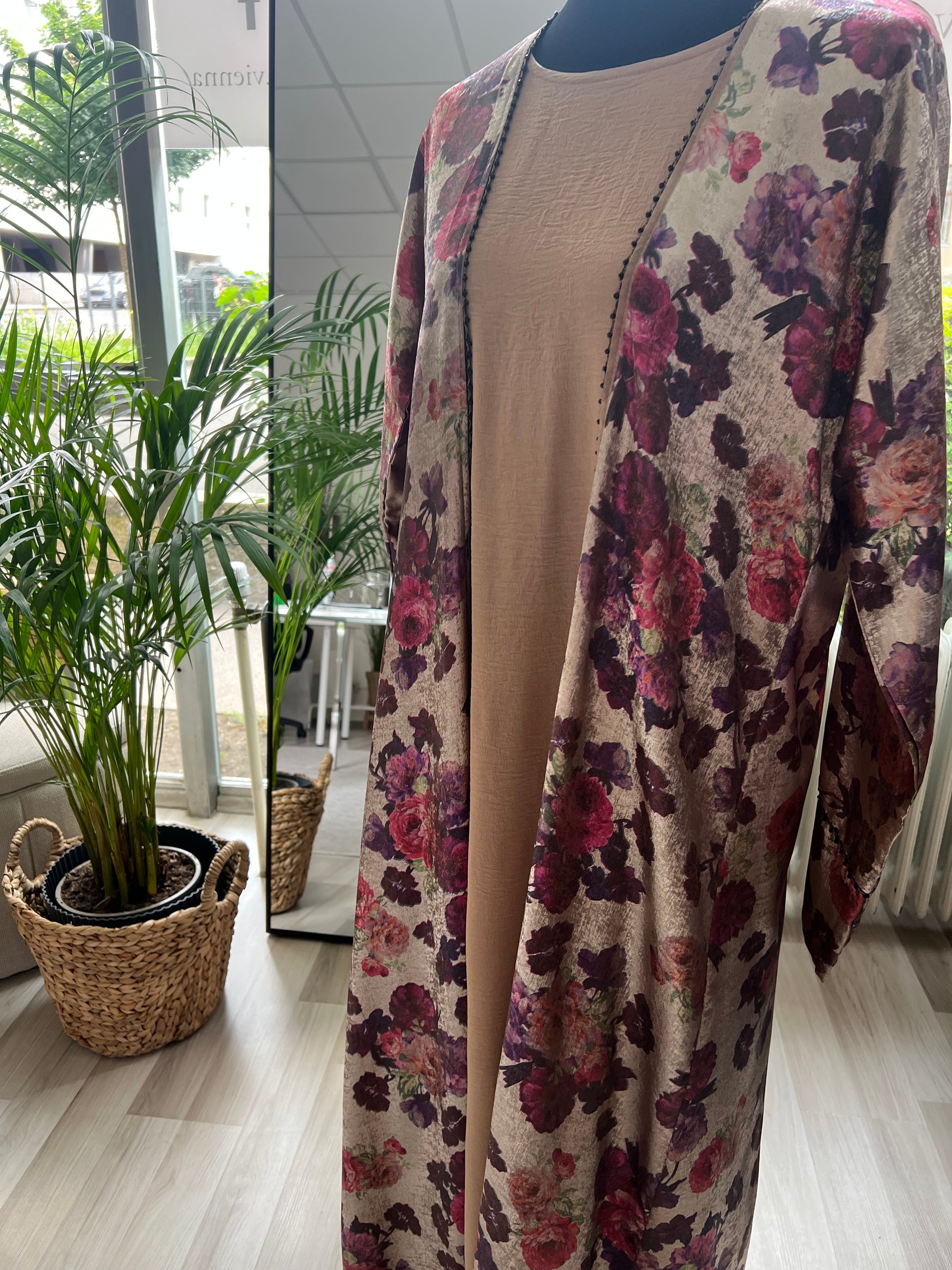 Seitliche Nahaufnahme der Abaya Rania in Blossom Gold vor einer Fensterfront mit Pflanzen – der florale Glanzstoff wirkt besonders edel im Tageslicht.“