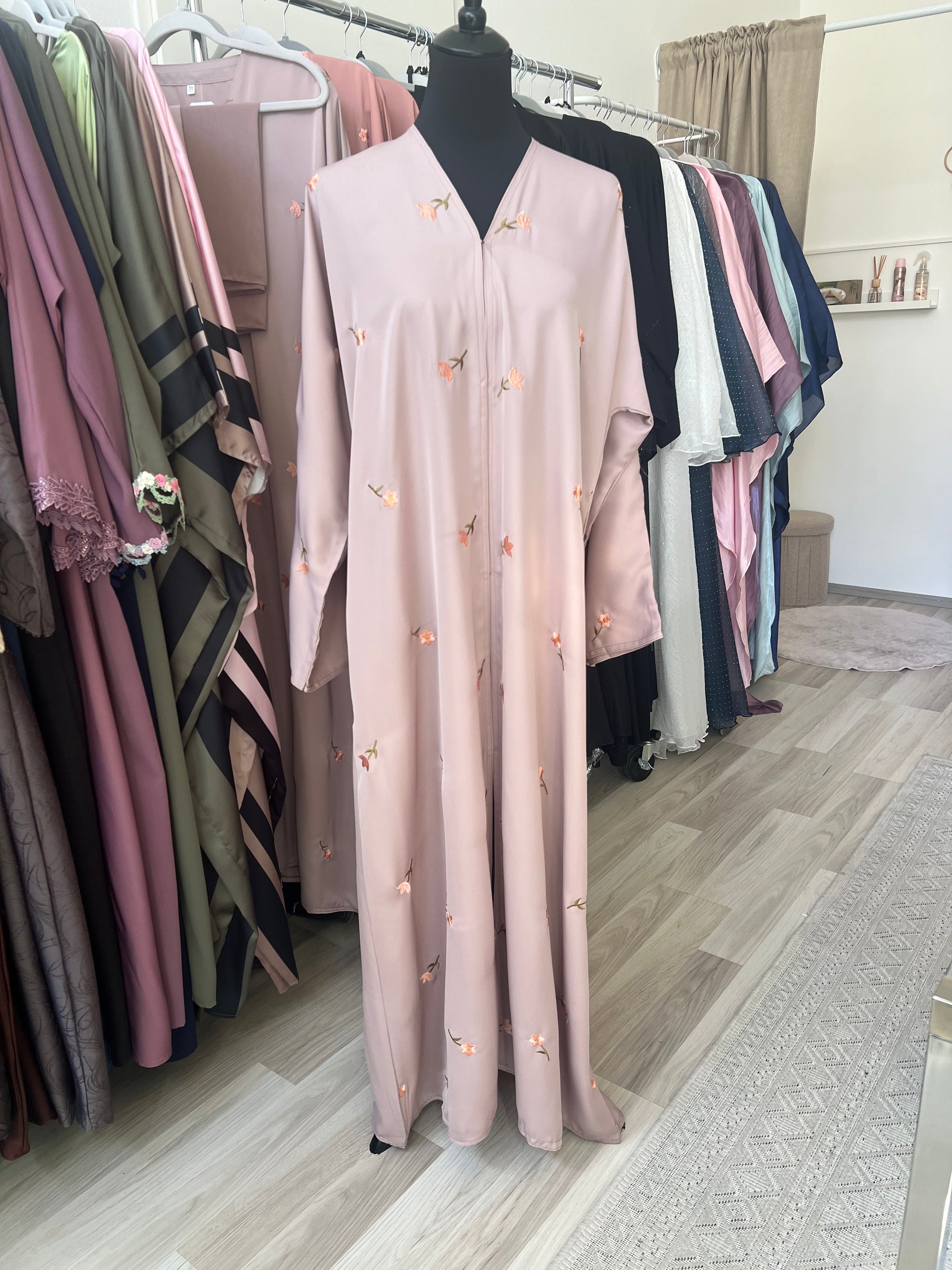 Beige Abaya mit floraler Stickerei auf Schneiderpuppe, umgeben von weiteren Abayas im Sheylin Showroom in Wien.