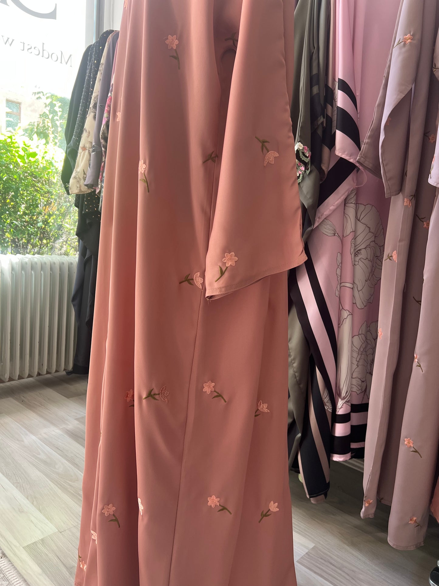 Seitliche Ansicht der pfirsichfarbenen Abaya mit feiner floraler Stickerei – fotografiert bei natürlichem Tageslicht im Showroom.