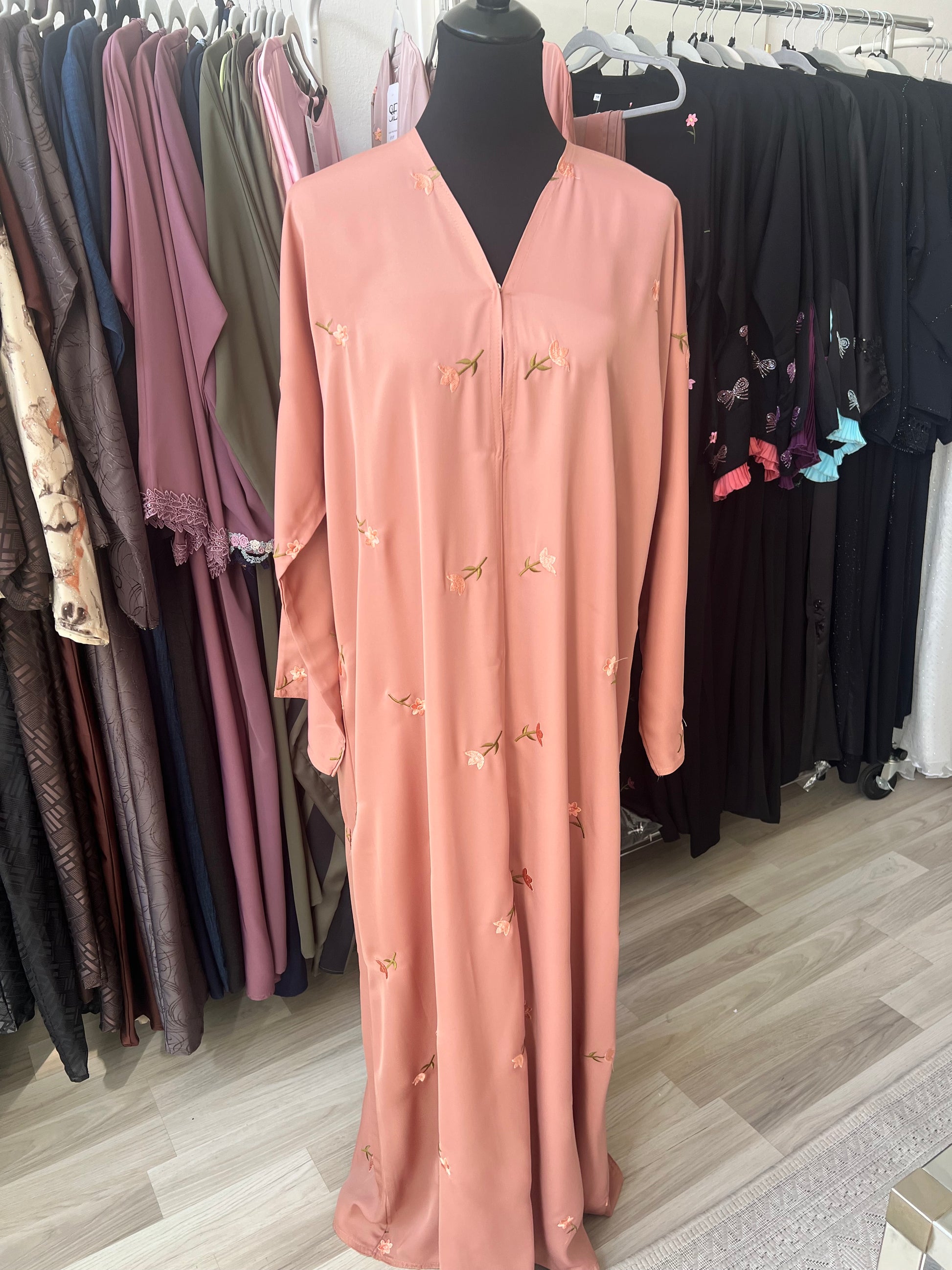 Pfirsichfarbene Abaya mit zarter Blumenstickerei, ausgestellt auf einer Schneiderpuppe im Sheylin Showroom, umgeben von weiteren Abayas.