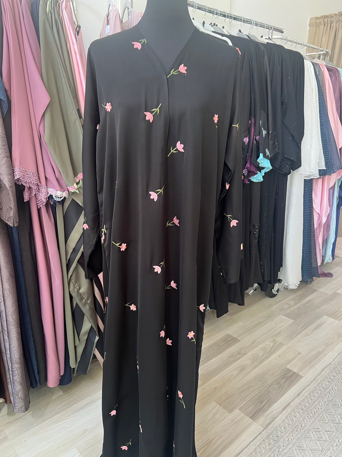 Schwarze Abaya mit pinker Blumenstickerei, elegant drapiert auf einer Schneiderpuppe im Sheylin Showroom zwischen weiteren Modellen.