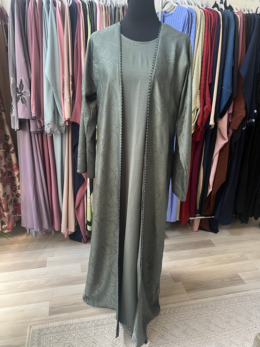 „Abaya Nura in Olivegrün – Komplettansicht mit Unterkleid, aufgenommen im Showroom vor Kleiderständern“